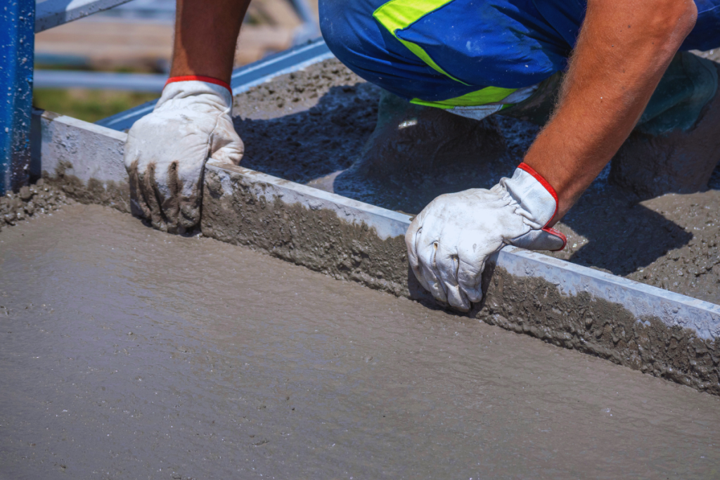 Close-up of gloved hands smoothing wet concrete along a metal form at a construction site, with blue work pants nearby.